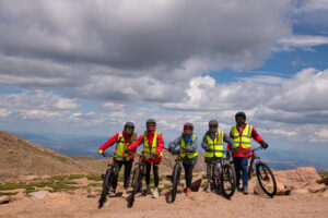 5 bike riders standing on rocks with clouds and mountains behind them